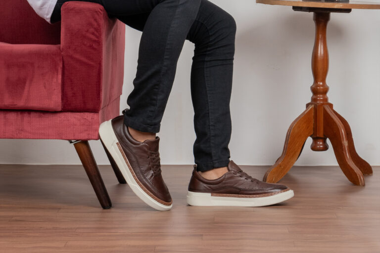 Imagem de um homem sentado, mostrando apenas do joelho para baixo, dando destaque para o sapatênis de couro legítimo em seus pés.